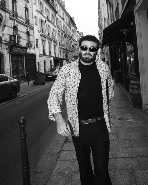 Black and white street-style portrait of a man with sunglasses in an open patterned shirt on a Parisian street.