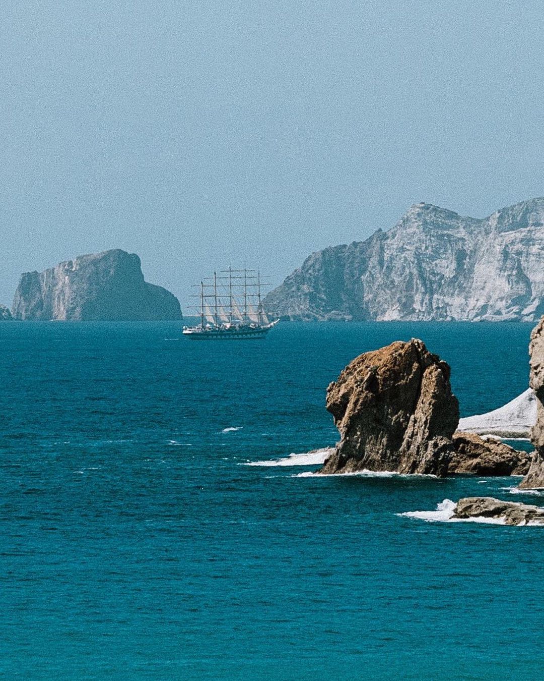 Tall ship sailing on blue ocean water near rocky coastline with island mountains in the distance under a clear sky.