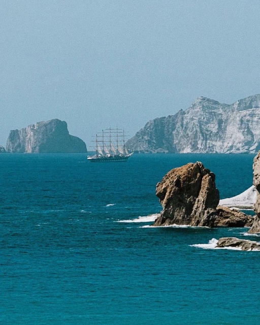 Tall ship sailing on blue ocean water near rocky coastline with island mountains in the distance under a clear sky.