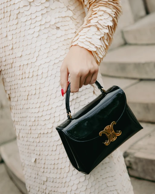 Adult female presents a black patent leather handbag with a gold logo, paired with a champagne sequin dress and red manicure.