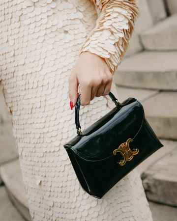 Adult female presents a black patent leather handbag with a gold logo, paired with a champagne sequin dress and red manicure.