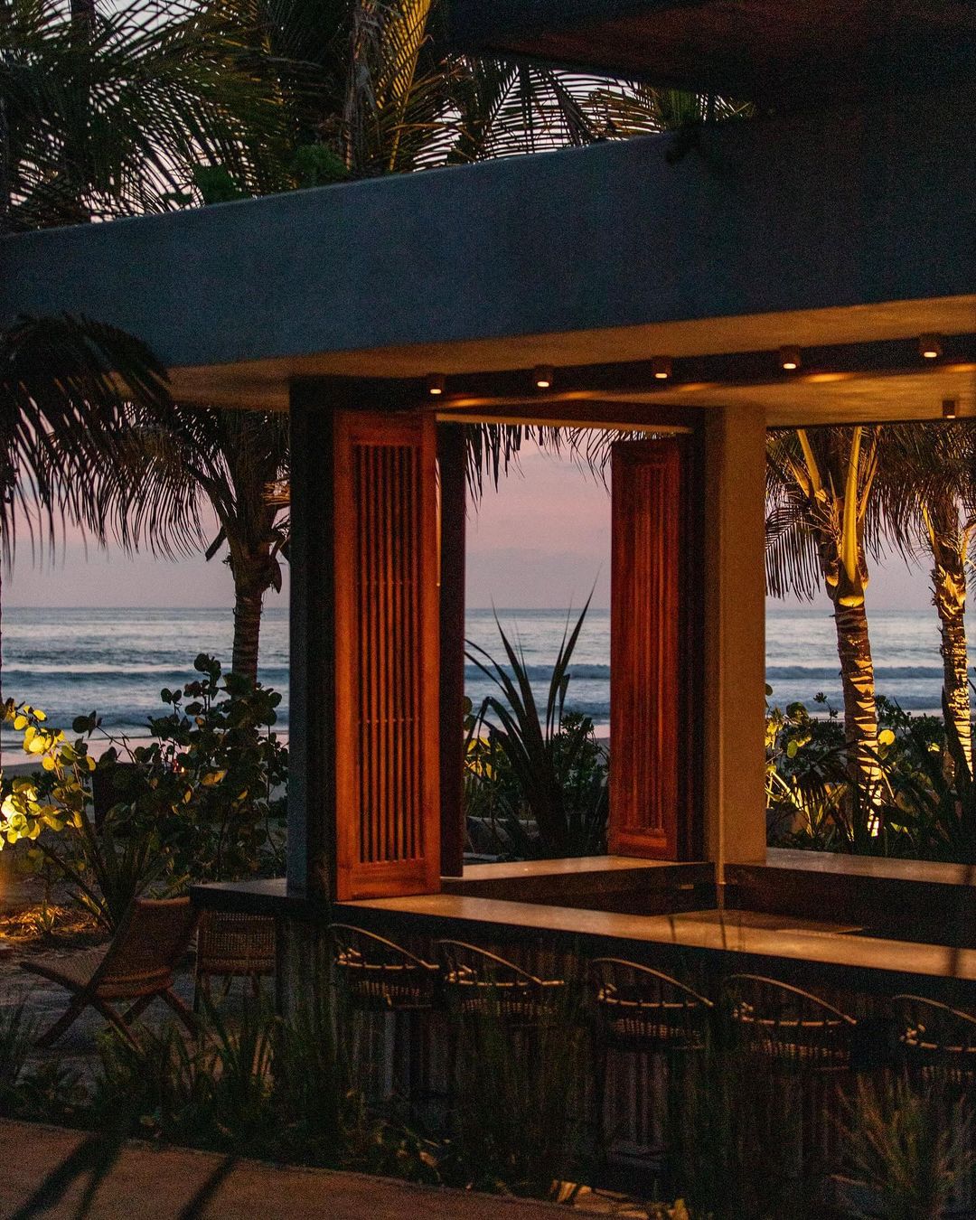 Open-air bar on the beach at sunset featuring wooden barstools and a view of the ocean, palm trees, and the sky.