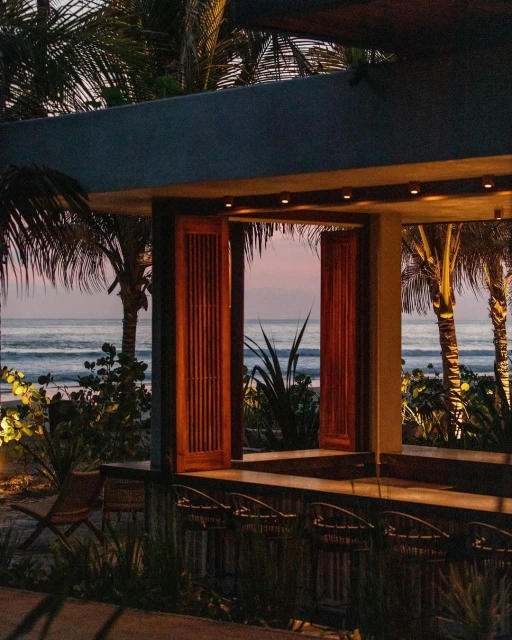 Open-air bar on the beach at sunset featuring wooden barstools and a view of the ocean, palm trees, and the sky.