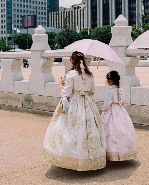 Two females in hanboks, traditional Korean dresses, walking in an urban setting with neutral stone bridge and tall buildings behind.