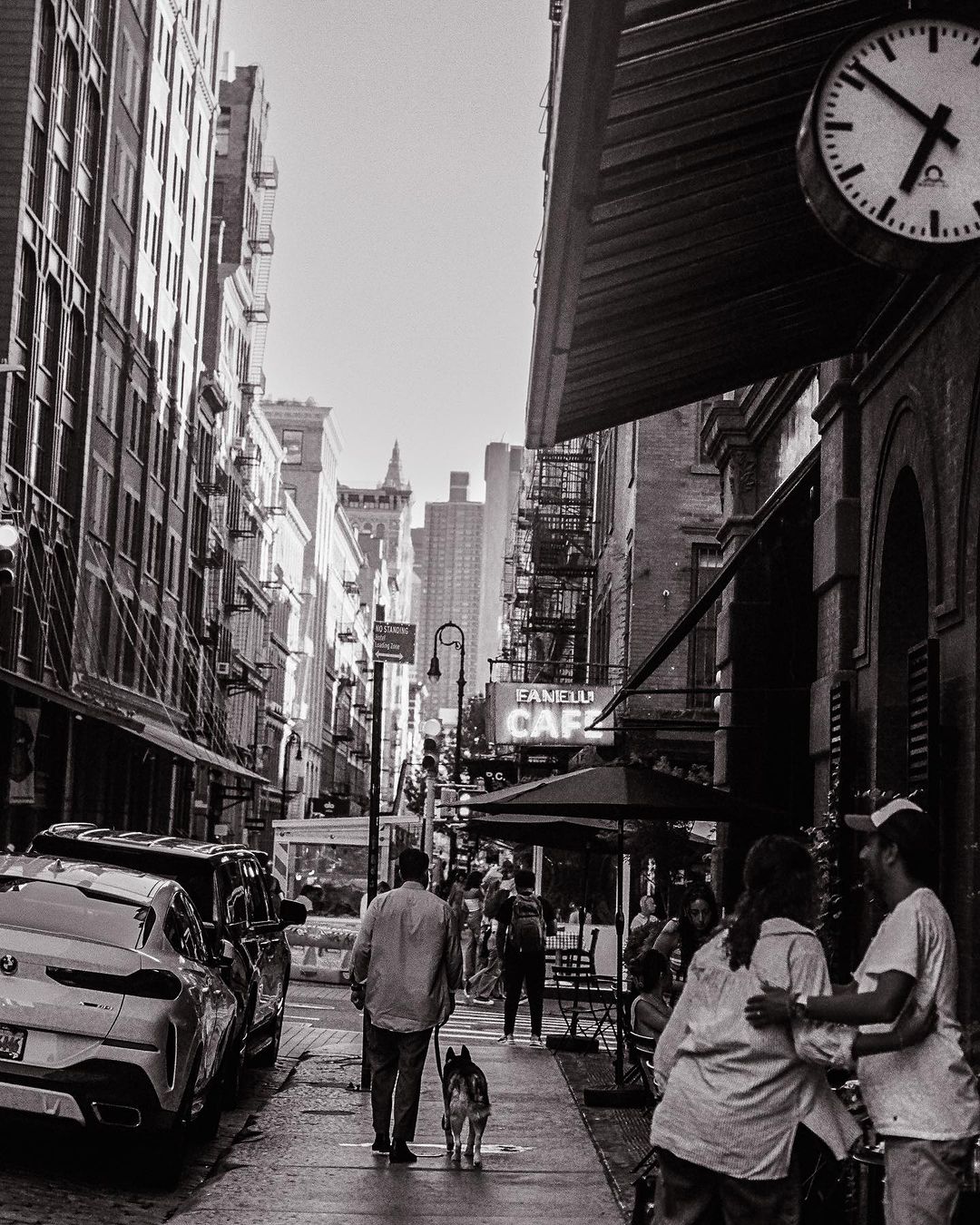 Black and white street scene with people at an outdoor cafe, a man walking a dog, parked cars, and tall buildings.