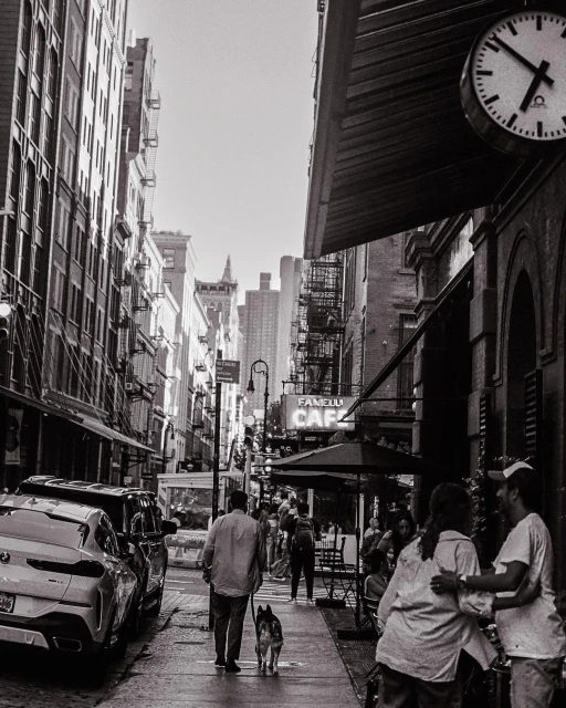 Black and white street scene with people at an outdoor cafe, a man walking a dog, parked cars, and tall buildings.