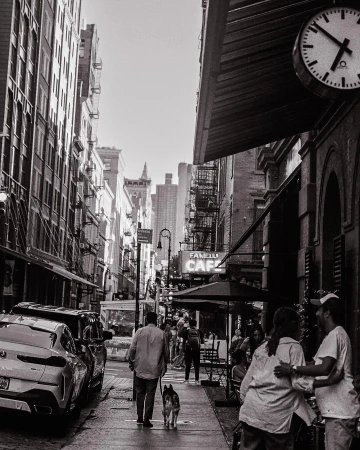 Black and white street scene with people at an outdoor cafe, a man walking a dog, parked cars, and tall buildings.