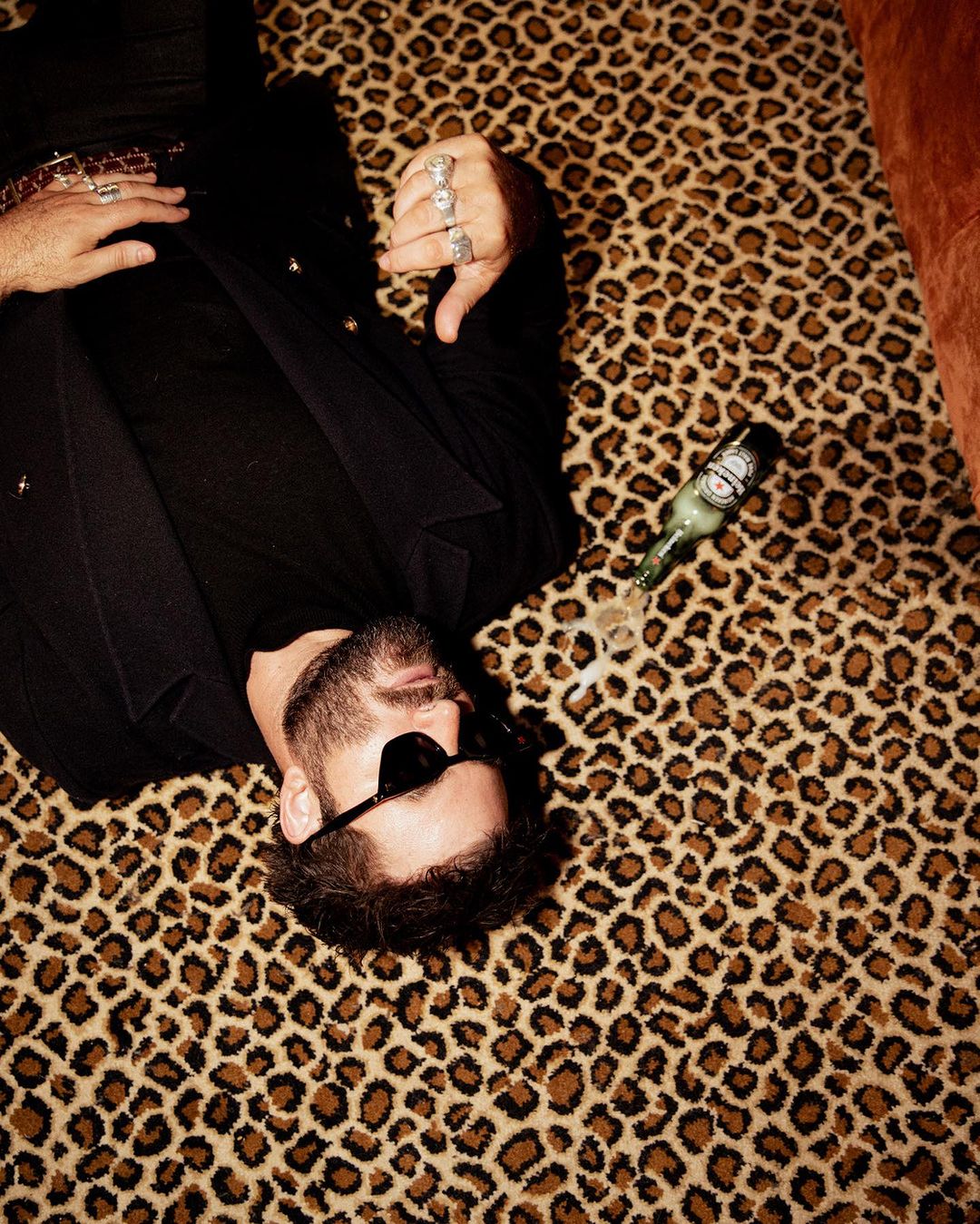 Adult male with sunglasses lies on an animal print rug next to an open green beer bottle in an indoor setting with bright light.