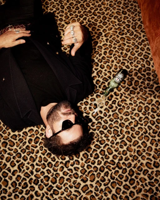 Adult male with sunglasses lies on an animal print rug next to an open green beer bottle in an indoor setting with bright light.