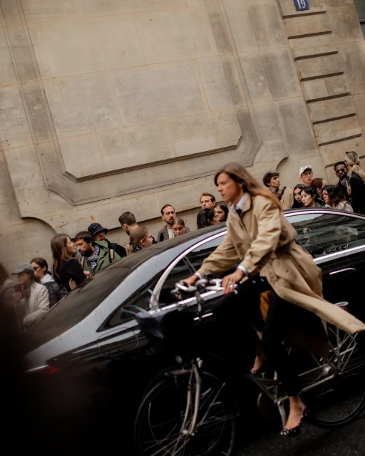 Woman in trench coat rides bicycle next to black car near crowd of people on urban street next to building with corner number.
