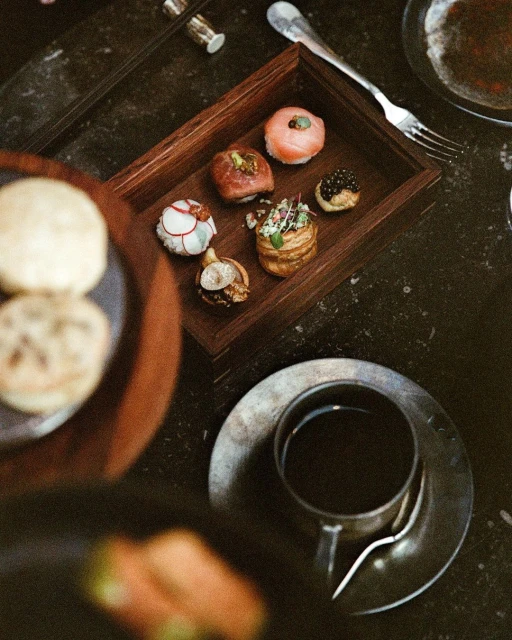 Overhead shot of assorted sushi in a wooden tray with coffee and silverware on a dark marble surface, with soft, natural lighting.
