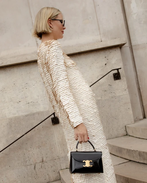A blonde woman in a cream dress with scale-like details and a black patent handbag stands on stone steps with a metal railing.