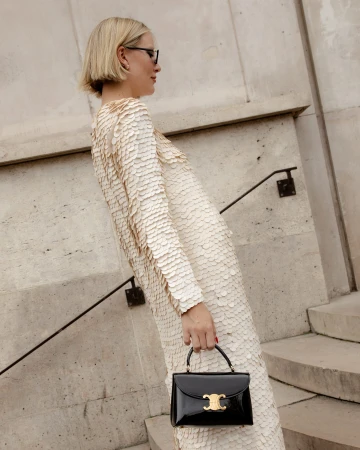 A blonde woman in a cream dress with scale-like details and a black patent handbag stands on stone steps with a metal railing.