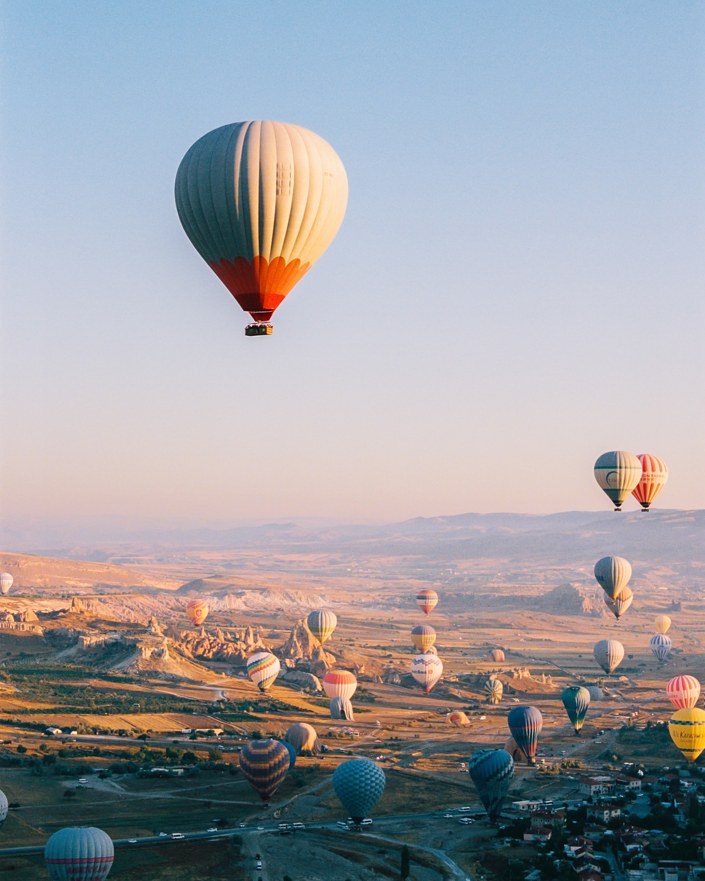 Hot air balloons float over a landscape at sunrise, creating a scenic view of Cappadocia, Turkey on a clear, sunny day.