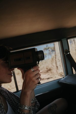 Adult woman filming through a vintage Super 8 camera inside of a car, medium shot, with a rural scene visible through the window.