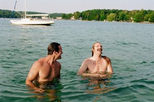 Two adult males are swimming in a lake with a sailboat in the background on a sunny day. They are shirtless and smiling.