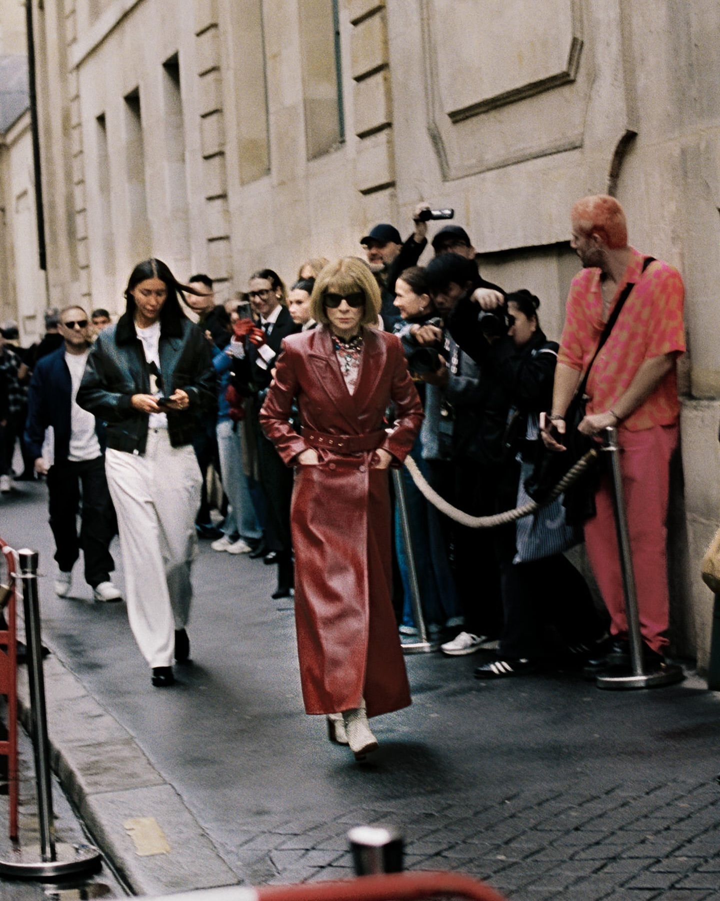 Fashion week scene featuring Anna Wintour in a long red coat walking past photographers and onlookers gathered outside a building.