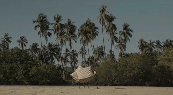 Wicker chair with striped pillows sits on a sandy beach beneath many tall palm trees under a clear sky in a tropical setting.