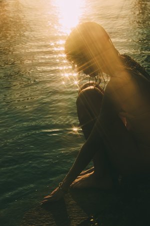 Woman sitting in water near a sandy beach with sunlight reflecting off the water's surface, creating a serene, contemplative scene.
