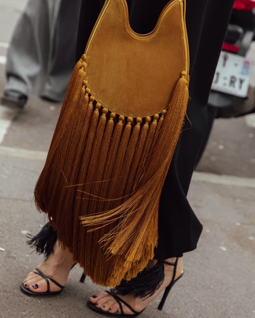 A woman's legs and feet are visible in black stiletto sandals that have black fringe on the ankle strap, along with a brown fringed bag.
