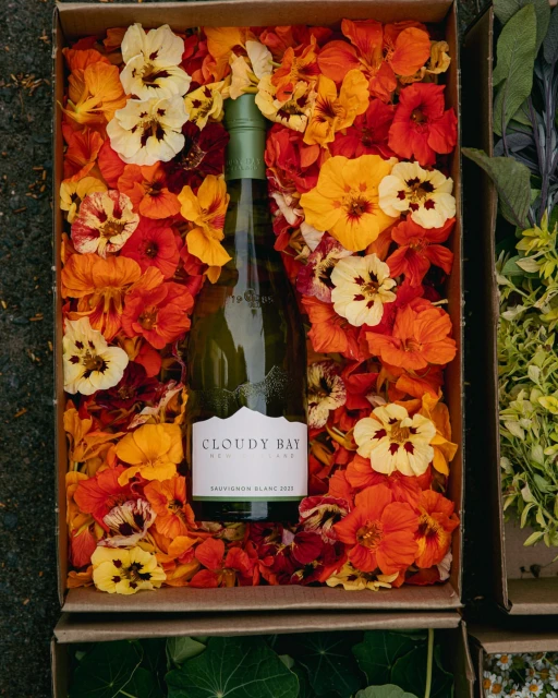 Bottle of Cloudy Bay Sauvignon Blanc wine nestled in a cardboard box filled with colorful orange and yellow nasturtium flowers.