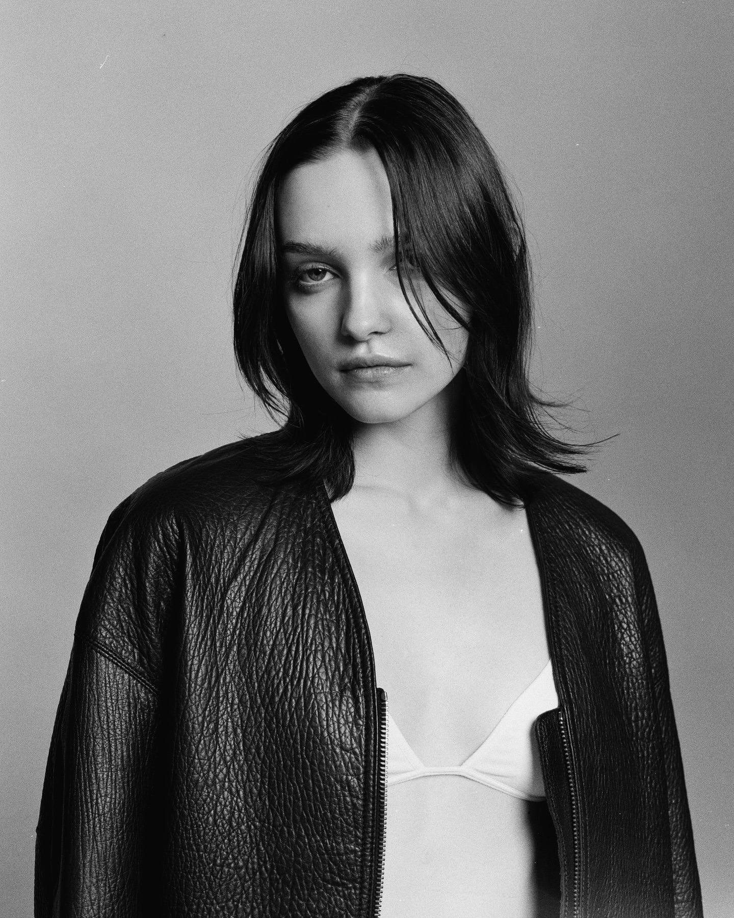 Monochrome portrait of a young adult woman with dark hair wearing a dark, zip-front leather jacket in front of a neutral background.