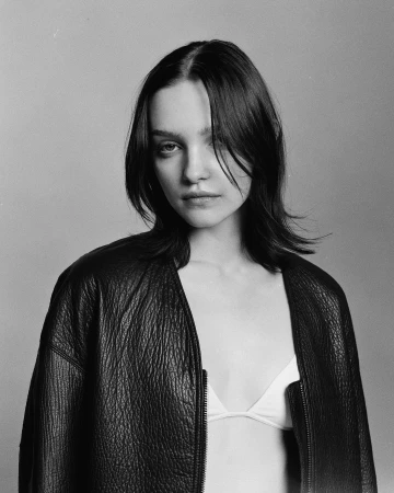 Monochrome portrait of a young adult woman with dark hair wearing a dark, zip-front leather jacket in front of a neutral background.