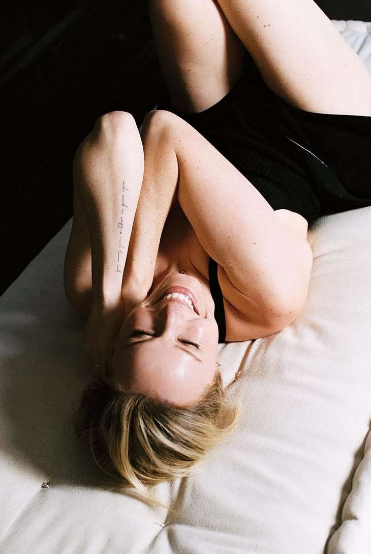 Woman lying on a cushion with her arms raised above her head, smiling, with blonde hair and a visible arm tattoo in a dimly lit space.