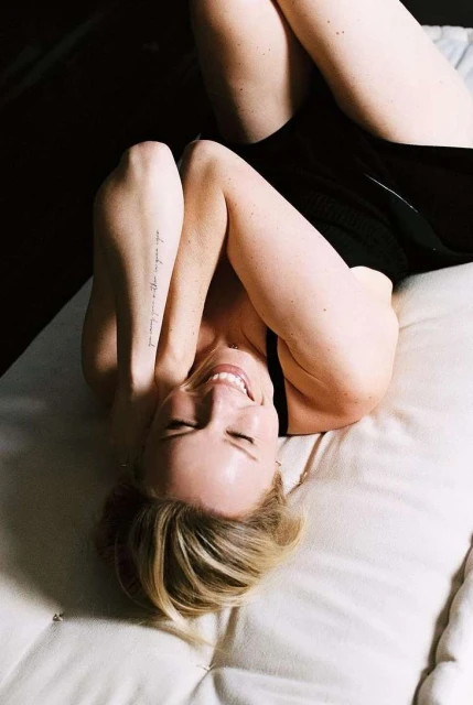 Woman lying on a cushion with her arms raised above her head, smiling, with blonde hair and a visible arm tattoo in a dimly lit space.