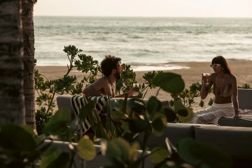 A man and a woman relaxing on a beach, enjoying drinks at sunset. Palm trees and ocean in background. Leisure, vacation, tropical setting.