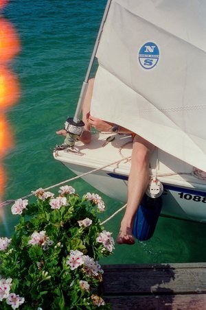 Sailboat is docked with a person's leg visible on deck and a flower plant next to the dock on a bright day with turquoise water.