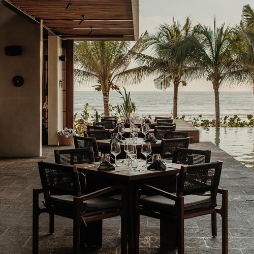 Outdoor restaurant setting with dark wood tables and chairs, set for dining with wine glasses and ocean view framed by palm trees.