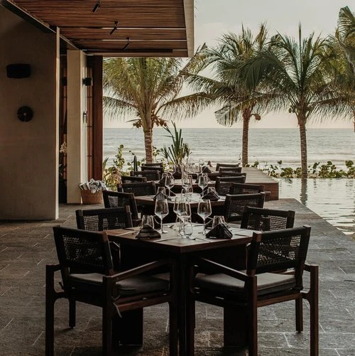 Outdoor restaurant setting with dark wood tables and chairs, set for dining with wine glasses and ocean view framed by palm trees.
