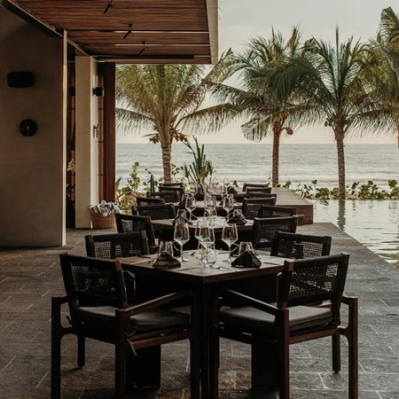 Outdoor restaurant setting with dark wood tables and chairs, set for dining with wine glasses and ocean view framed by palm trees.