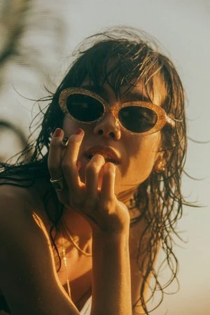 Close-up portrait of a woman with wet hair wearing glittery sunglasses and gold rings against a sunlit outdoor backdrop.
