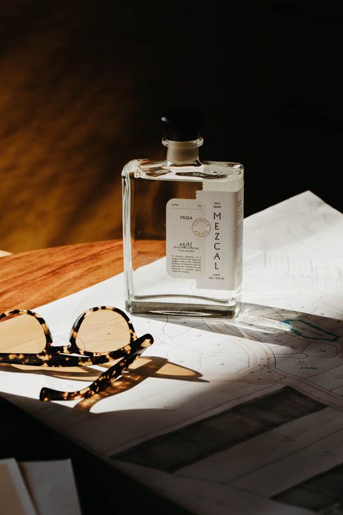 Still life photograph of a clear mezcal bottle with a black cap next to sunglasses on top of a nautical chart.