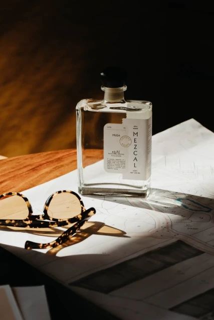 Still life photograph of a clear mezcal bottle with a black cap next to sunglasses on top of a nautical chart.