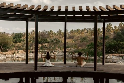 Two people practice yoga under a pergola overlooking a pond and tropical foliage in a serene, outdoor setting. Wellness theme.