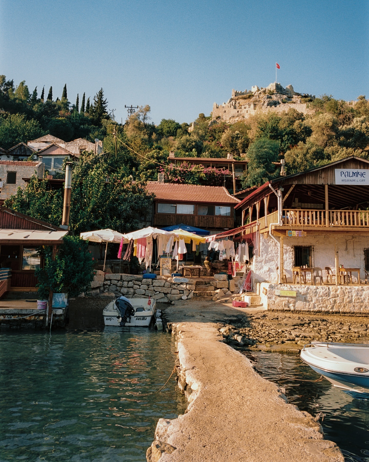 Scenic view of a Mediterranean village with traditional buildings, boats, and a stone pier along a coastline, under a clear blue sky.