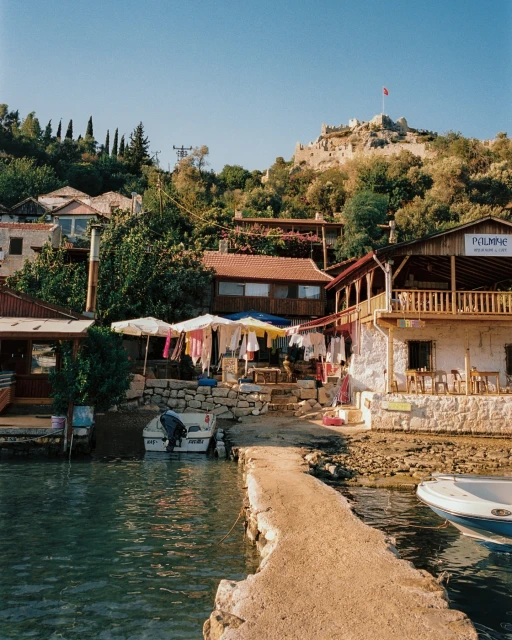 Scenic view of a Mediterranean village with traditional buildings, boats, and a stone pier along a coastline, under a clear blue sky.