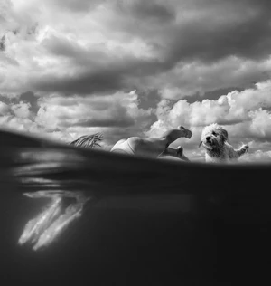 A woman lies in the water next to a fluffy white dog in a black and white photograph with a cloudy sky backdrop.