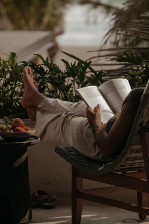 Person relaxing on a wicker chair reading an open book with feet up, beside a bowl of fresh fruit.