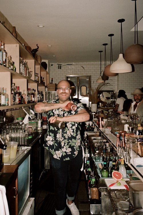 Bald adult male bartender in a Hawaiian shirt mixing drinks in a shaker behind a bar with bottles and customers in an indoor setting.