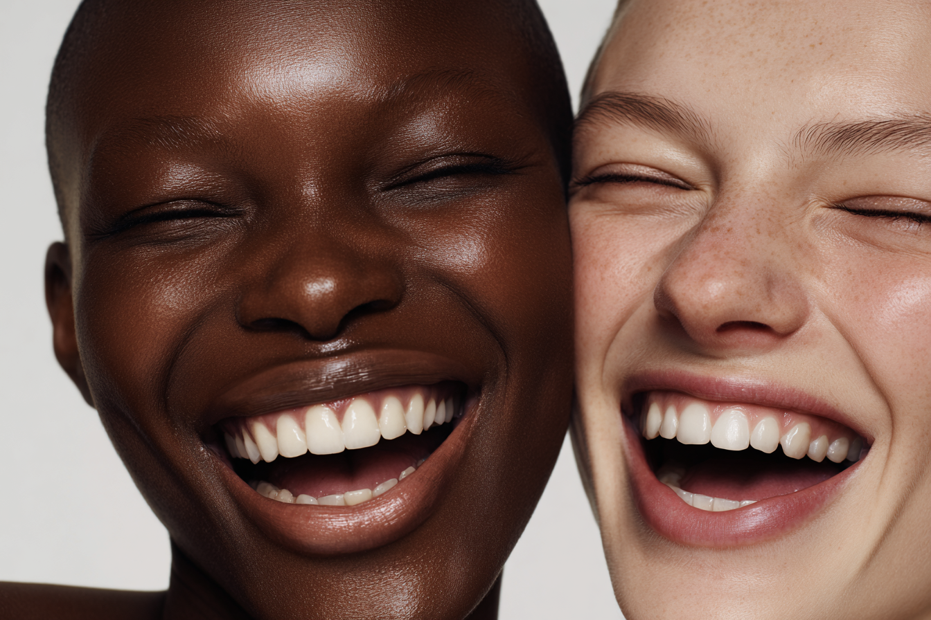 Close-up portrait of two adult women, one African American and one Caucasian, laughing with their eyes closed in a beauty shot.