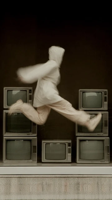 A person in white clothing jumps over a stack of old televisions against a dark background, creating a dynamic and surreal visual.