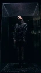 Woman in dark textured jacket standing inside a transparent glass cube covered in tiny water droplets.