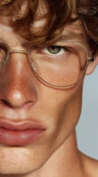 Close up portrait of a young man with curly brown hair wearing thin wire frame glasses and freckles against a neutral background