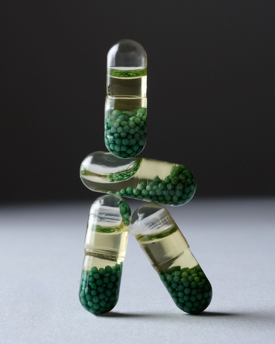 Still life shot of semi-transparent capsules with green spheres and liquid inside, stacked against a dark gray background.