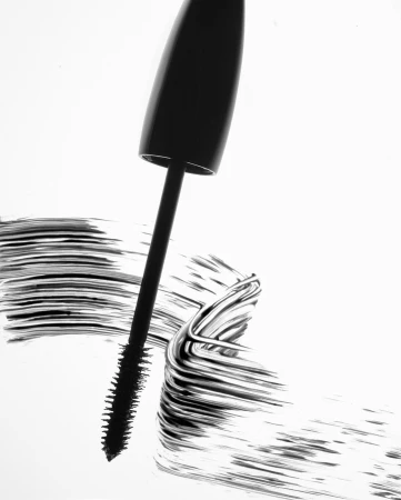 Close-up of a black mascara wand and tube creating a bold black makeup swatch on white paper against a bright white background.