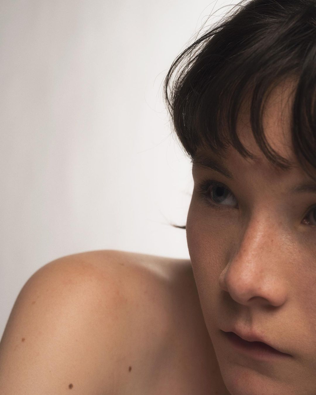 Close-up portrait of an adult woman with dark hair, looking off-camera in a soft, diffused light against a plain, light-colored backdrop.